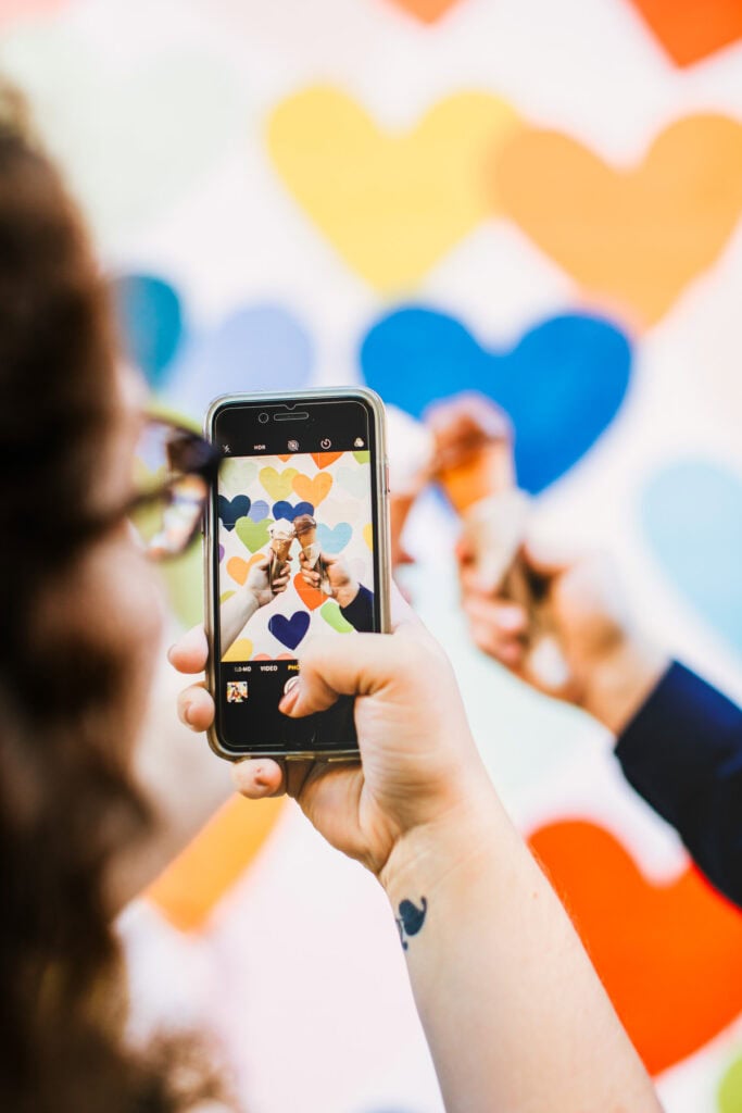 Susannah Brinkley Henry takes a photo with her phone of two people posing in front of a colorful wall covered with heart shapes.