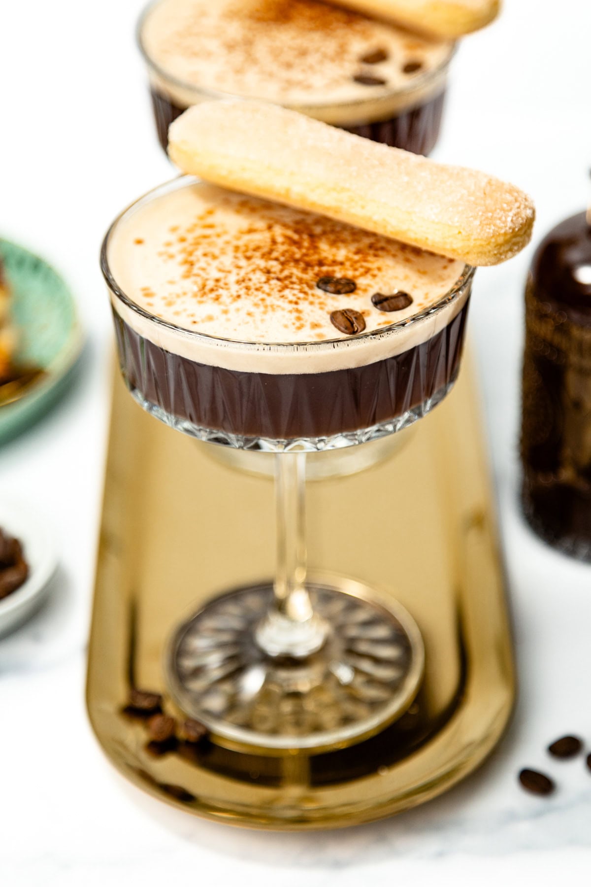 A glass of espresso martini topped with cocoa powder and coffee beans, garnished with a ladyfinger biscuit, sits on a gold tray.