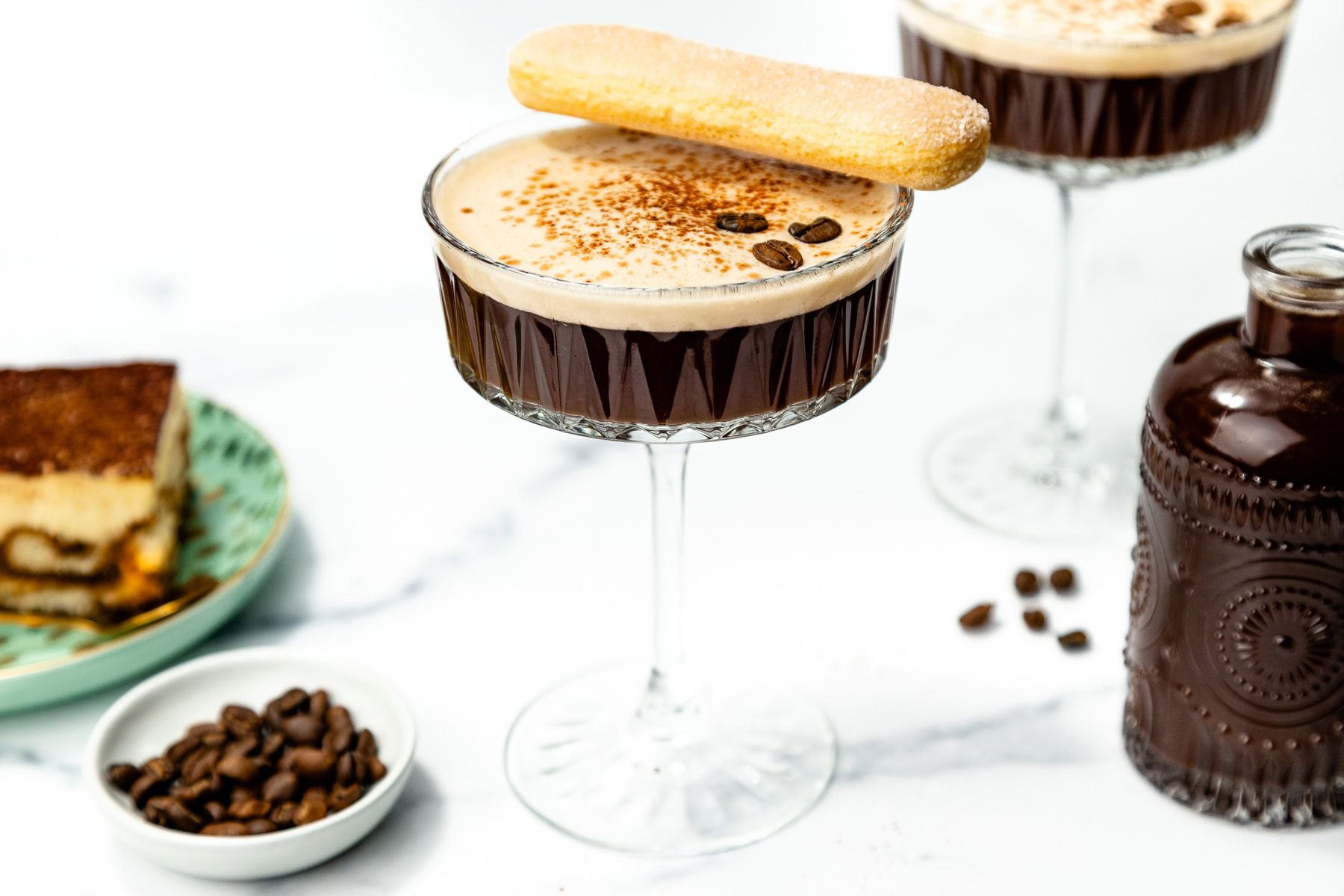 A glass of espresso martini topped with a ladyfinger biscuit and coffee beans, next to a small bottle, a plate of tiramisu, and a bowl of coffee beans on a white surface.