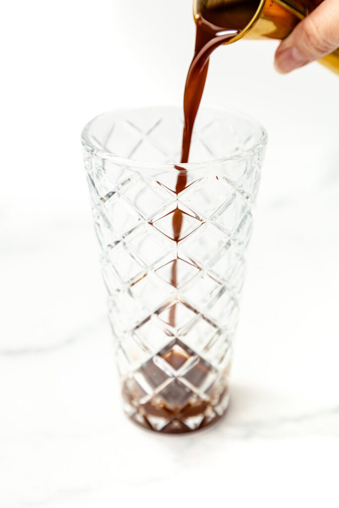 Coffee is being poured from a metal container into a transparent, patterned glass on a white surface.