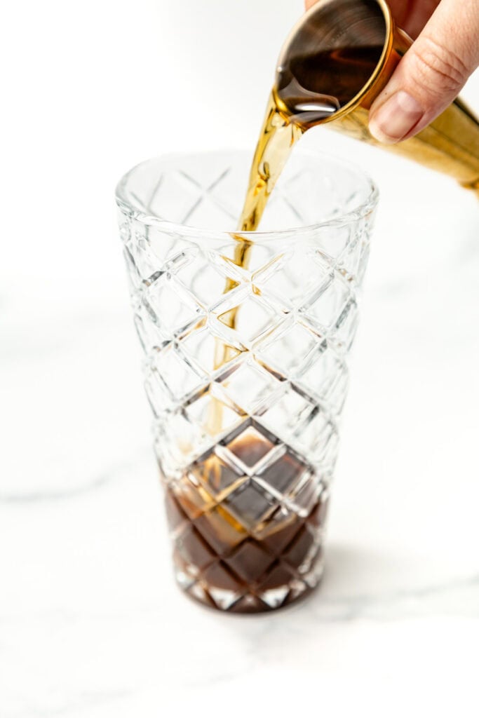 A hand pours a dark liquid from a measuring cup into a clear, patterned glass on a white surface.