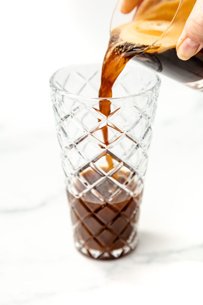 Coffee is being poured from a glass container into a tall, empty, patterned glass on a white surface.