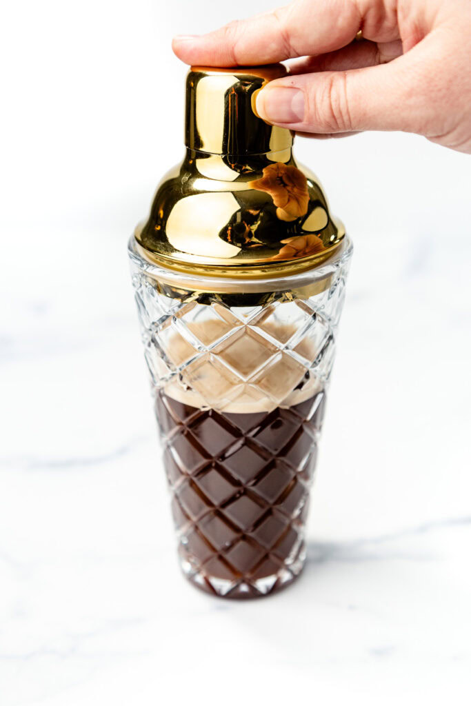 A hand holding a gold cocktail shaker with a patterned glass base, filled with a dark liquid, on a white surface.