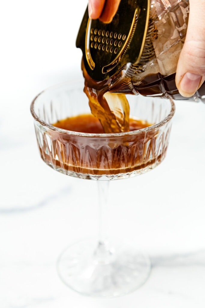 A hand pours a dark brown cocktail from a shaker into a coupe glass on a white marble surface.