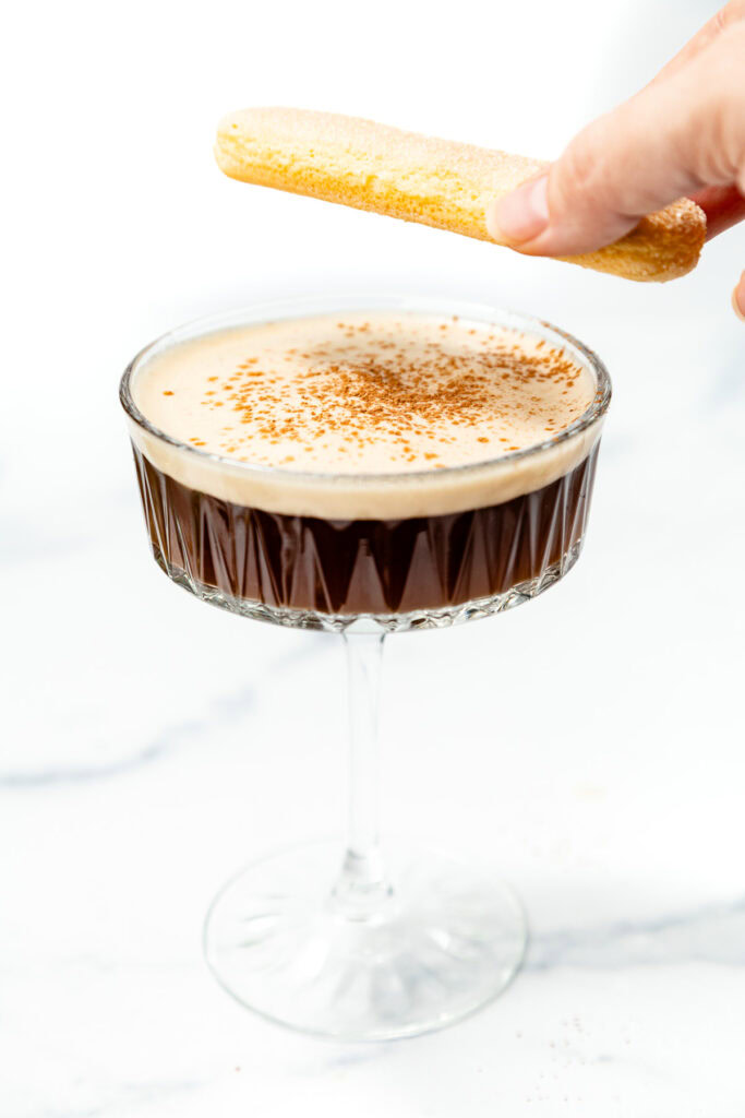 A hand holding a ladyfinger biscuit above a glass of espresso martini garnished with cocoa powder on a white surface.