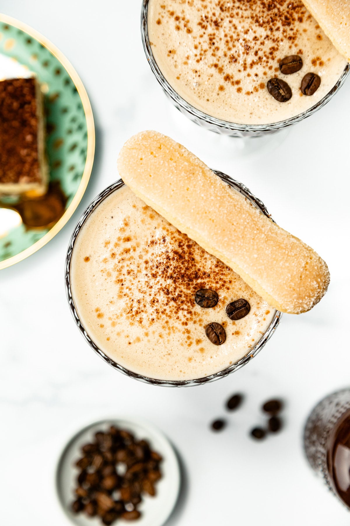 Overhead view of two creamy coffee drinks topped with cocoa powder, coffee beans, and a ladyfinger biscuit. A plate with dessert and a bowl of coffee beans are nearby.