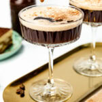 A hand dips a ladyfinger biscuit into a coupe glass filled with a frothy espresso martini, with another glass and dessert visible in the background.