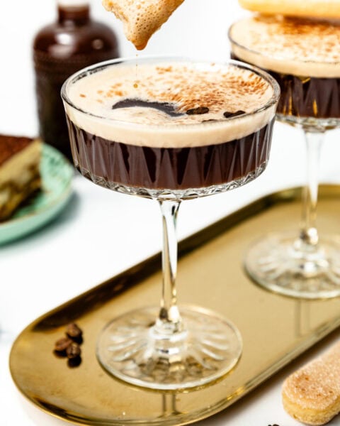 A hand dips a ladyfinger biscuit into a coupe glass filled with a frothy espresso martini, with another glass and dessert visible in the background.