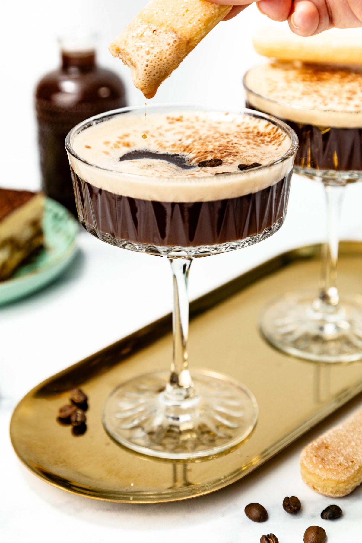 A hand dips a ladyfinger biscuit into a coupe glass filled with a frothy espresso martini, with another glass and dessert visible in the background.