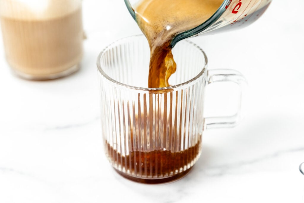 Coffee is being poured from a glass measuring cup into a clear, ribbed glass mug on a white surface.