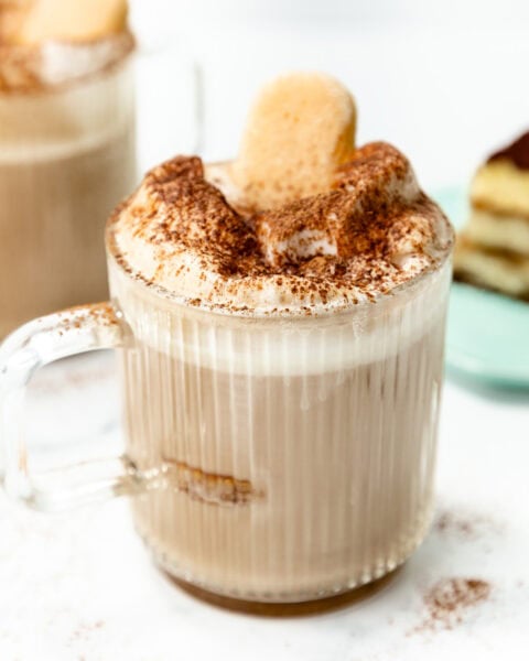 A glass mug filled with a frothy beverage topped with cocoa powder and a ladyfinger cookie, with a slice of dessert on a plate in the background.