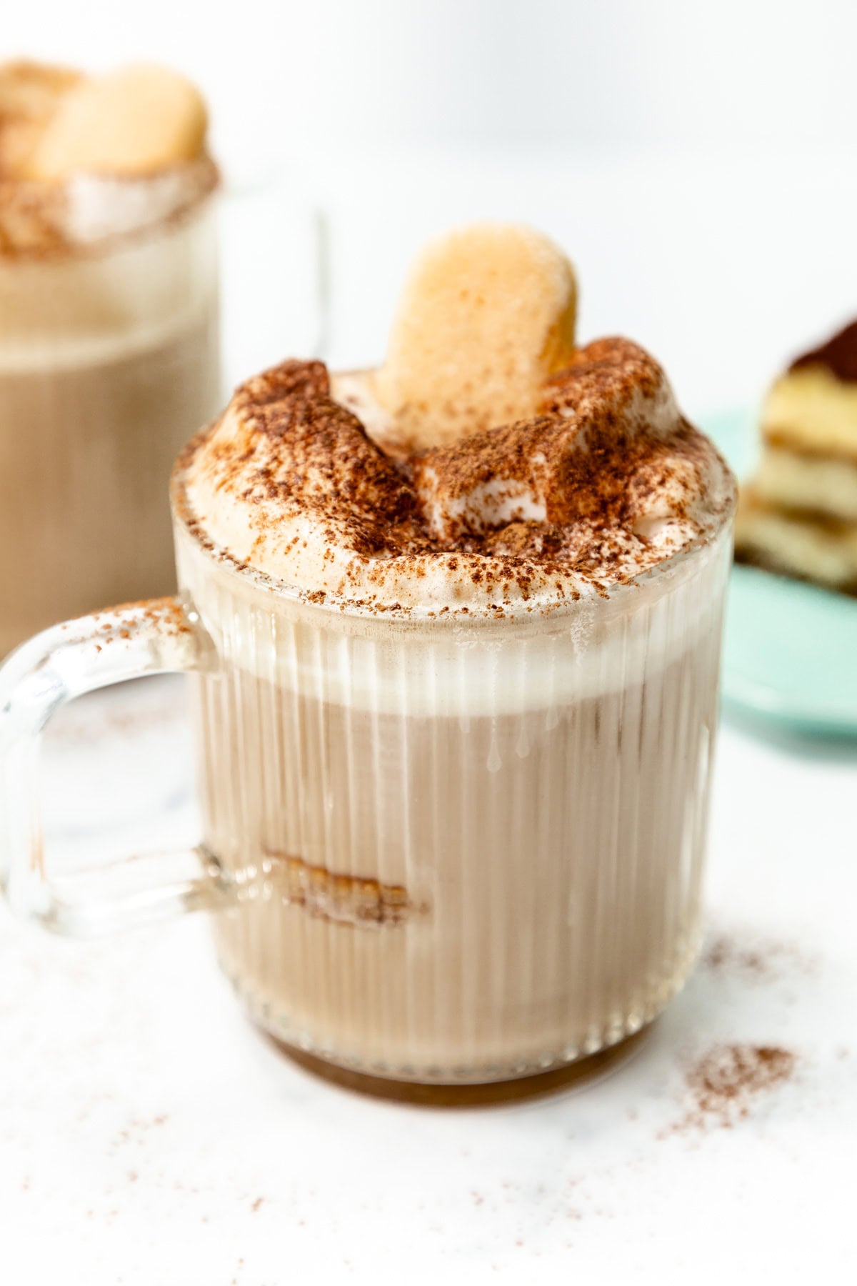 A glass mug filled with a frothy beverage topped with cocoa powder and a ladyfinger cookie, with a slice of dessert on a plate in the background.