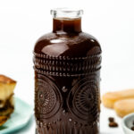 A clear glass bottle filled with dark liquid, likely coffee or liqueur, sits on a white surface beside dessert items and coffee beans.