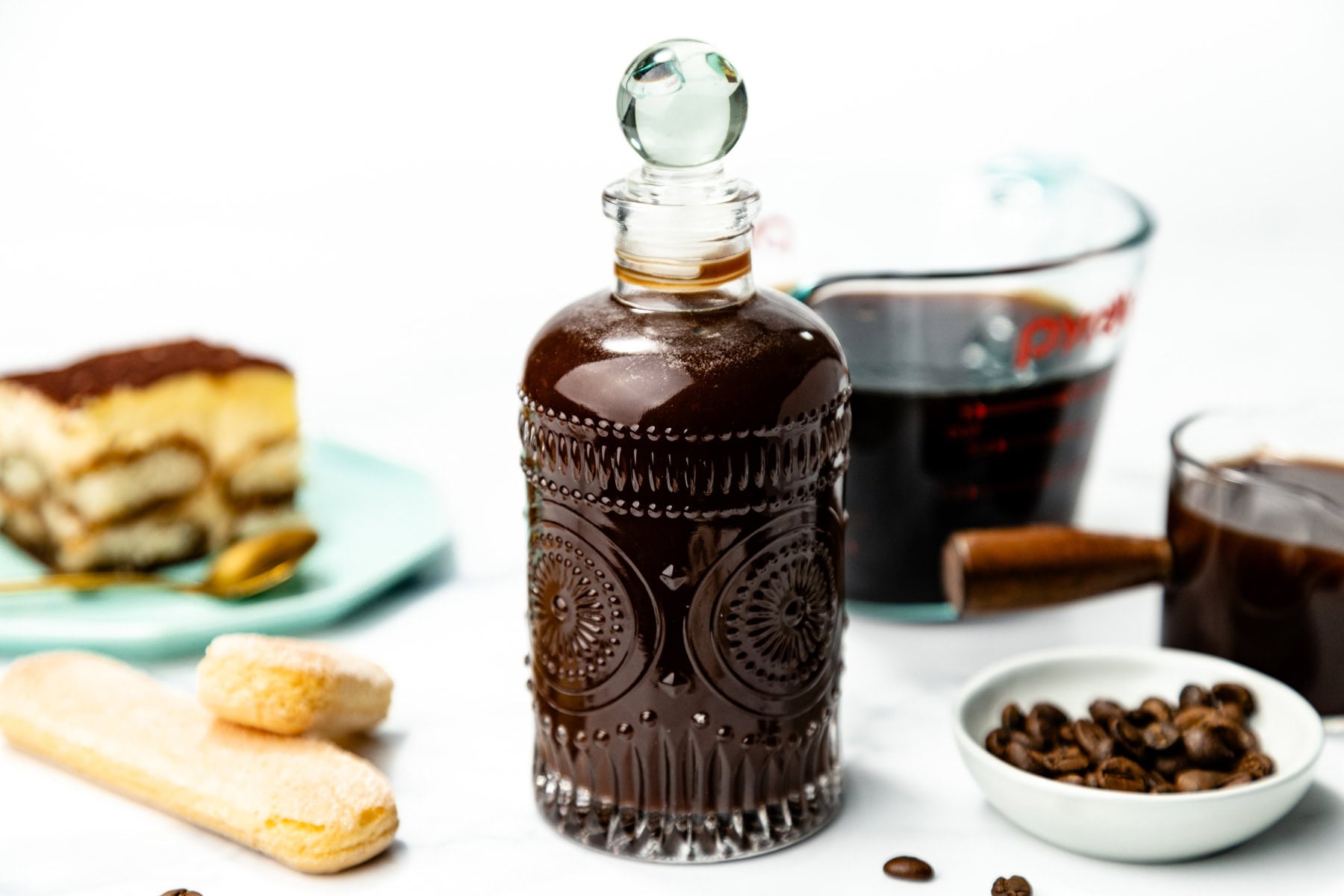 A decorative glass bottle filled with dark liquid sits on a counter with ladyfingers, coffee beans, a cup of coffee, and a slice of dessert in the background.