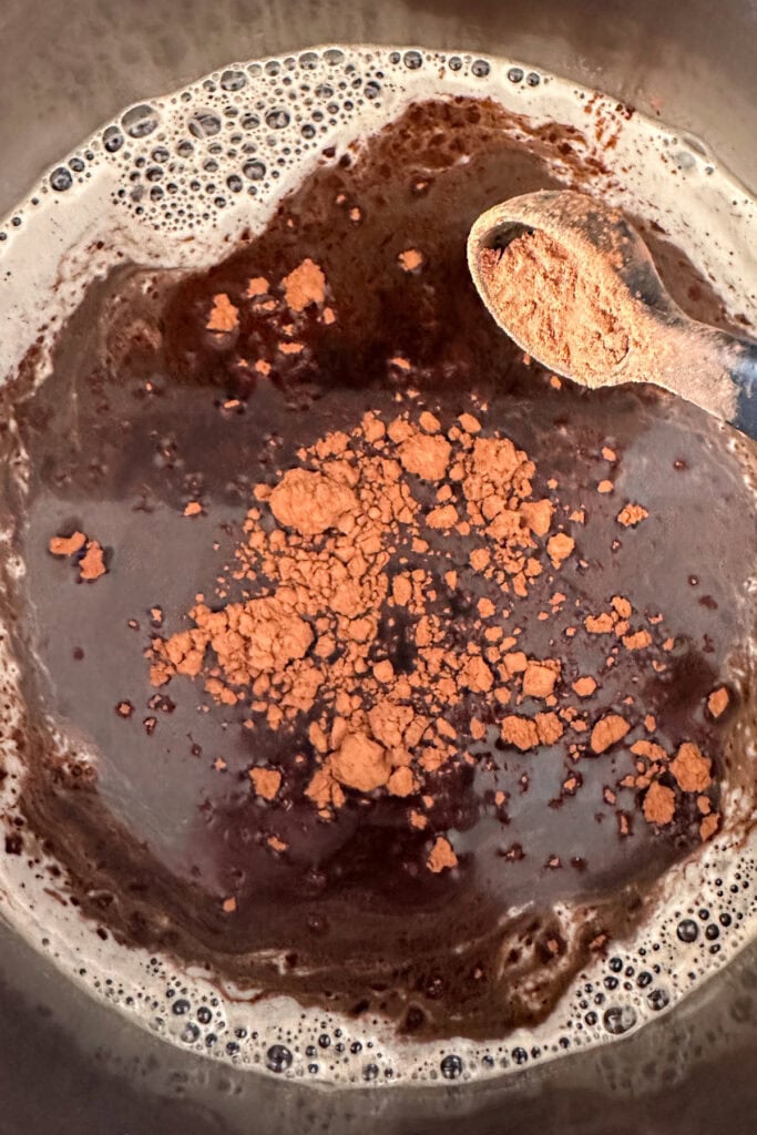 A close-up of cocoa powder being added to a pot of tiramisu syrup, with a wooden spoon holding more cocoa powder above the rich mixture.