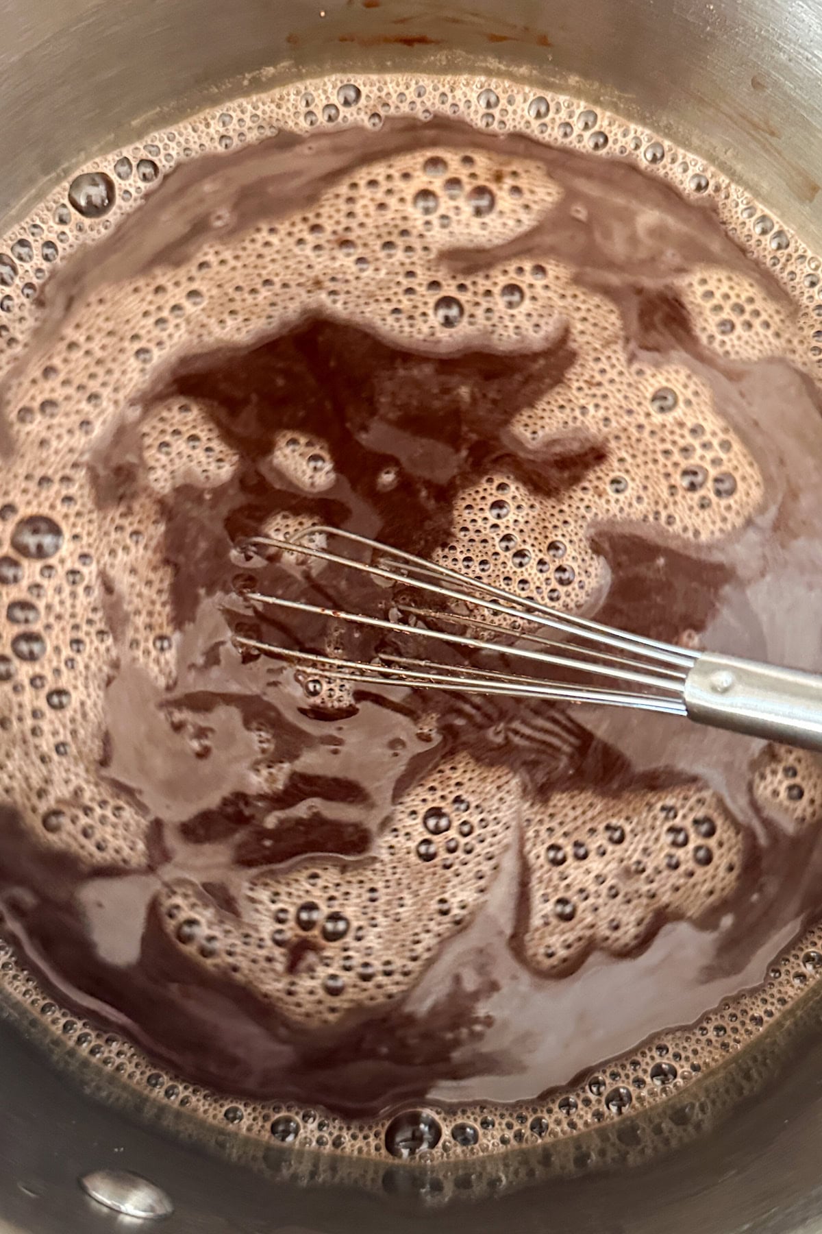 A metal whisk stirs a pot of foamy, dark brown liquid, likely tiramisu syrup or cocoa mixture, inside a stainless steel saucepan.