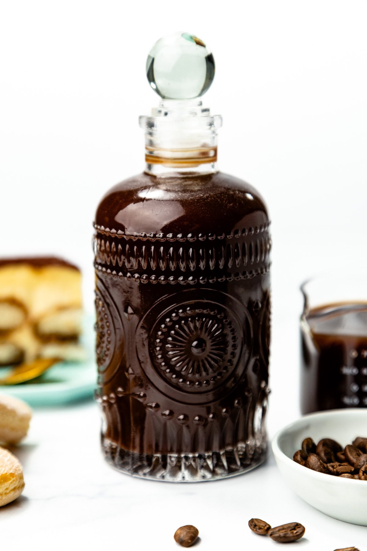A clear, decorative glass bottle filled with dark liquid, surrounded by a bowl of coffee beans and plates with dessert on a white surface.