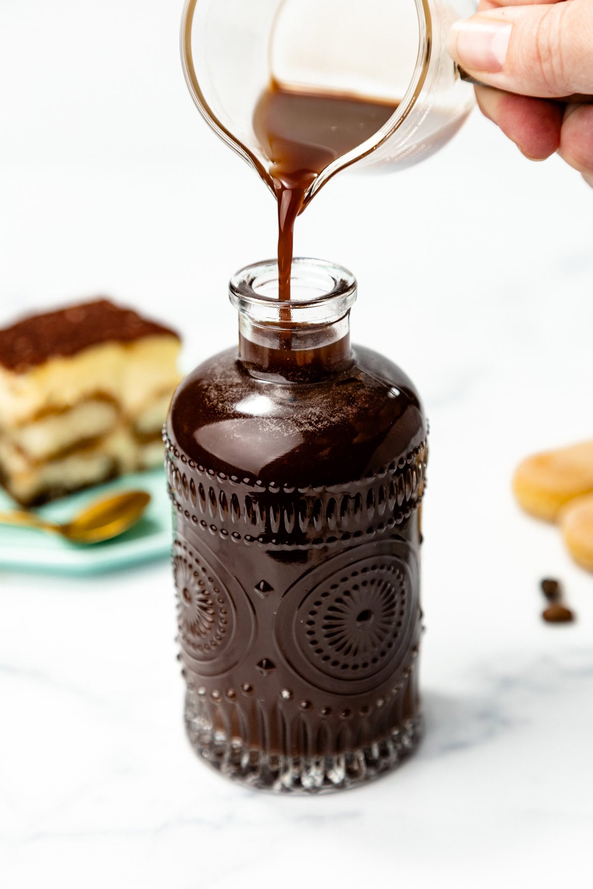 A hand pours dark liquid from a small pitcher into a decorative glass bottle, with a dessert and spoon in the background.