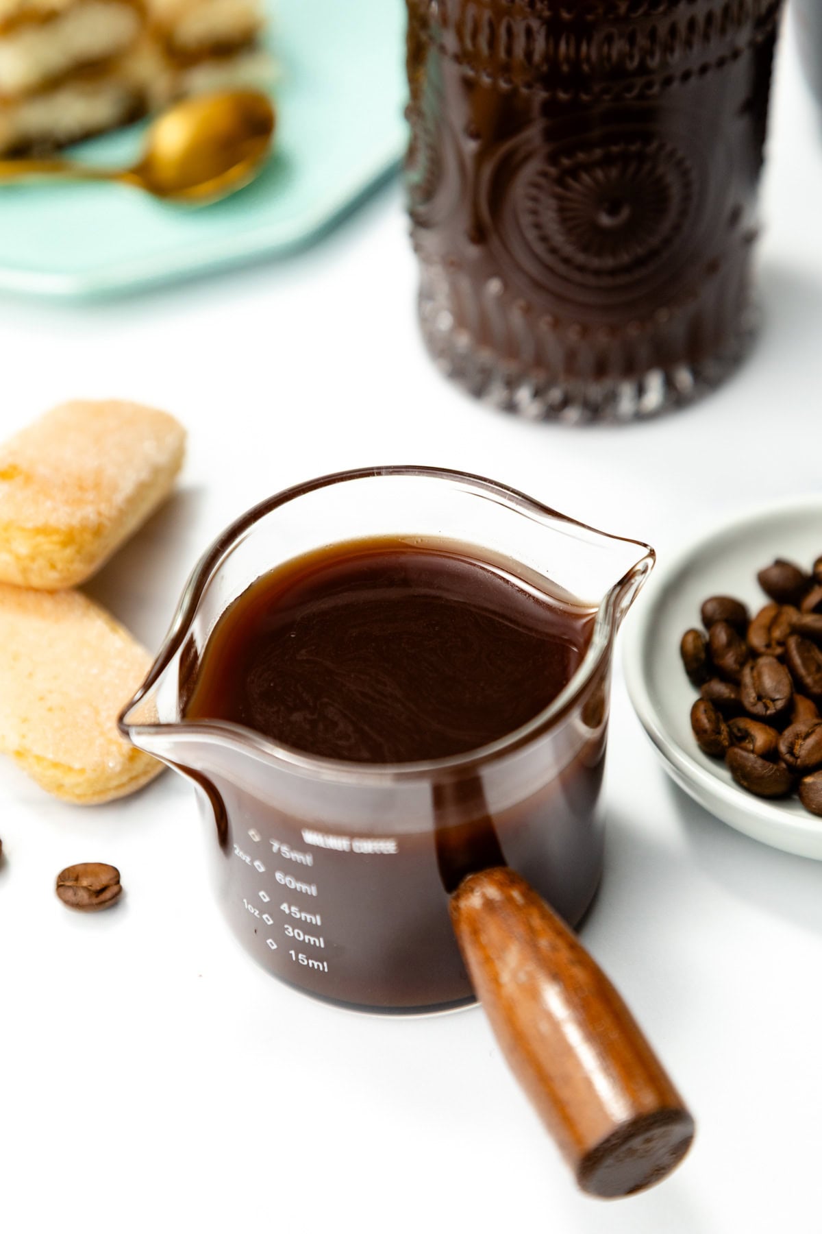A glass measuring cup filled with black coffee sits on a white surface, surrounded by coffee beans, ladyfinger biscuits, and a dark glass jar.
