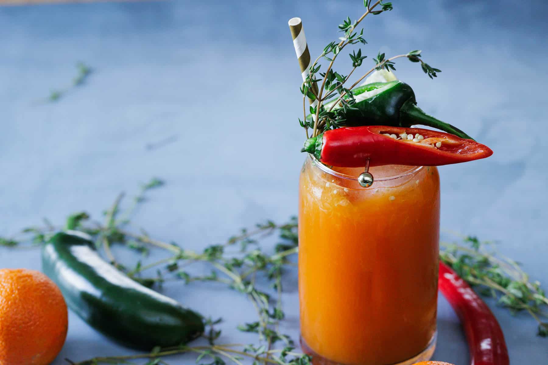 A glass of orange juice garnished with herbs and whole chili peppers, with a striped straw, sits on a blue surface next to fresh chilies and thyme.