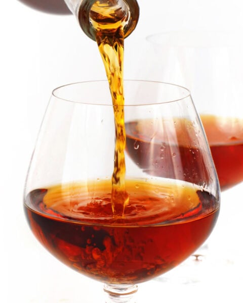 A close-up of brown liquid being poured from a bottle into a clear, round glass, with another similar glass in the background.