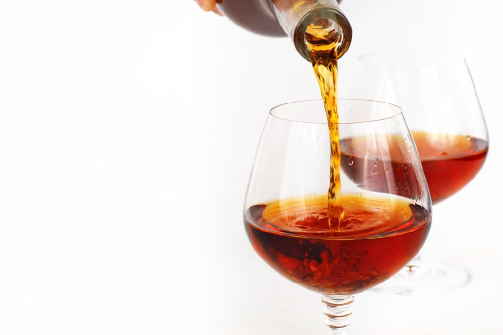 A bottle pours amber liquid into a round glass, with another filled glass in the background, both on a white surface.