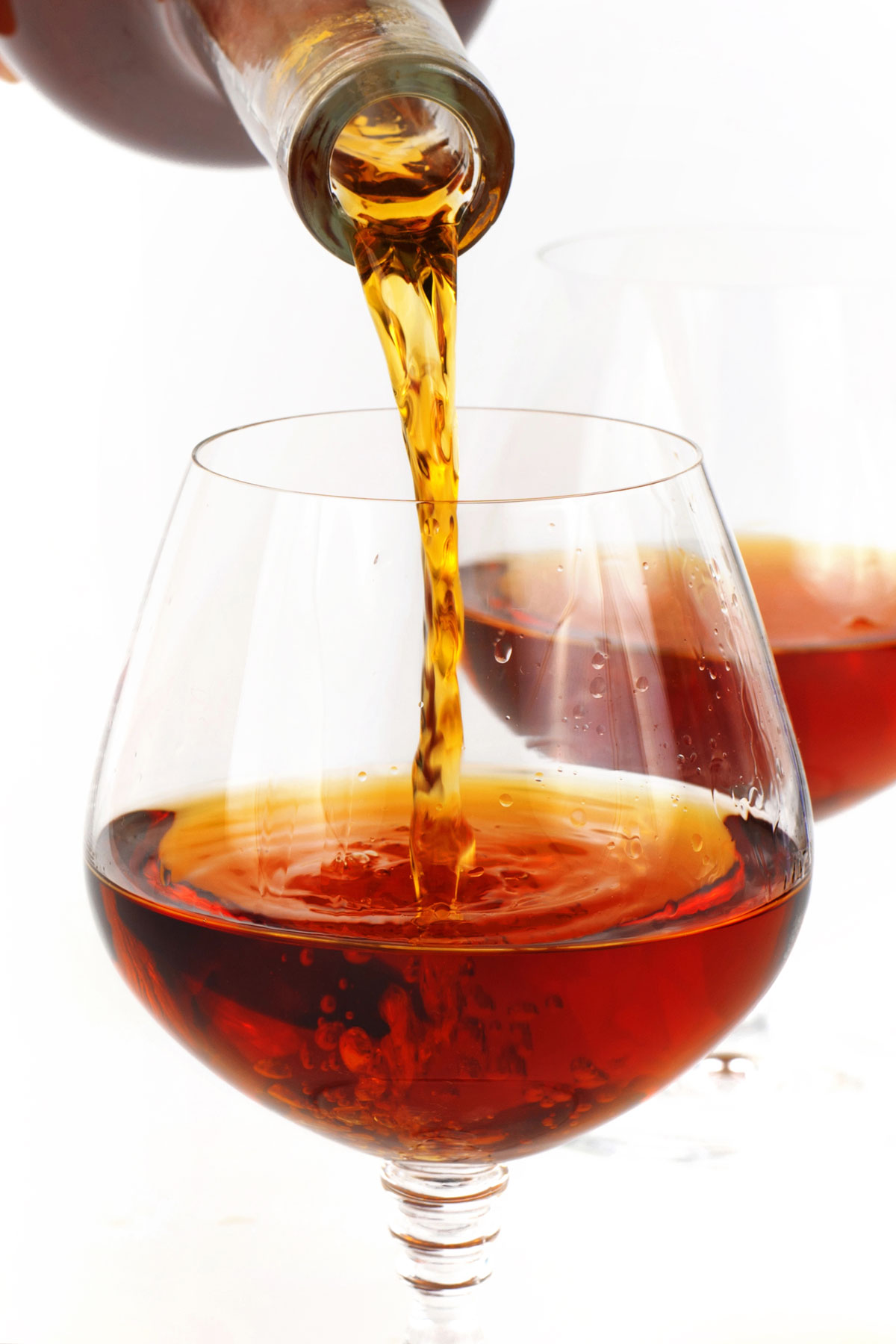 A close-up of brown liquid being poured from a bottle into a clear, round glass, with another similar glass in the background.