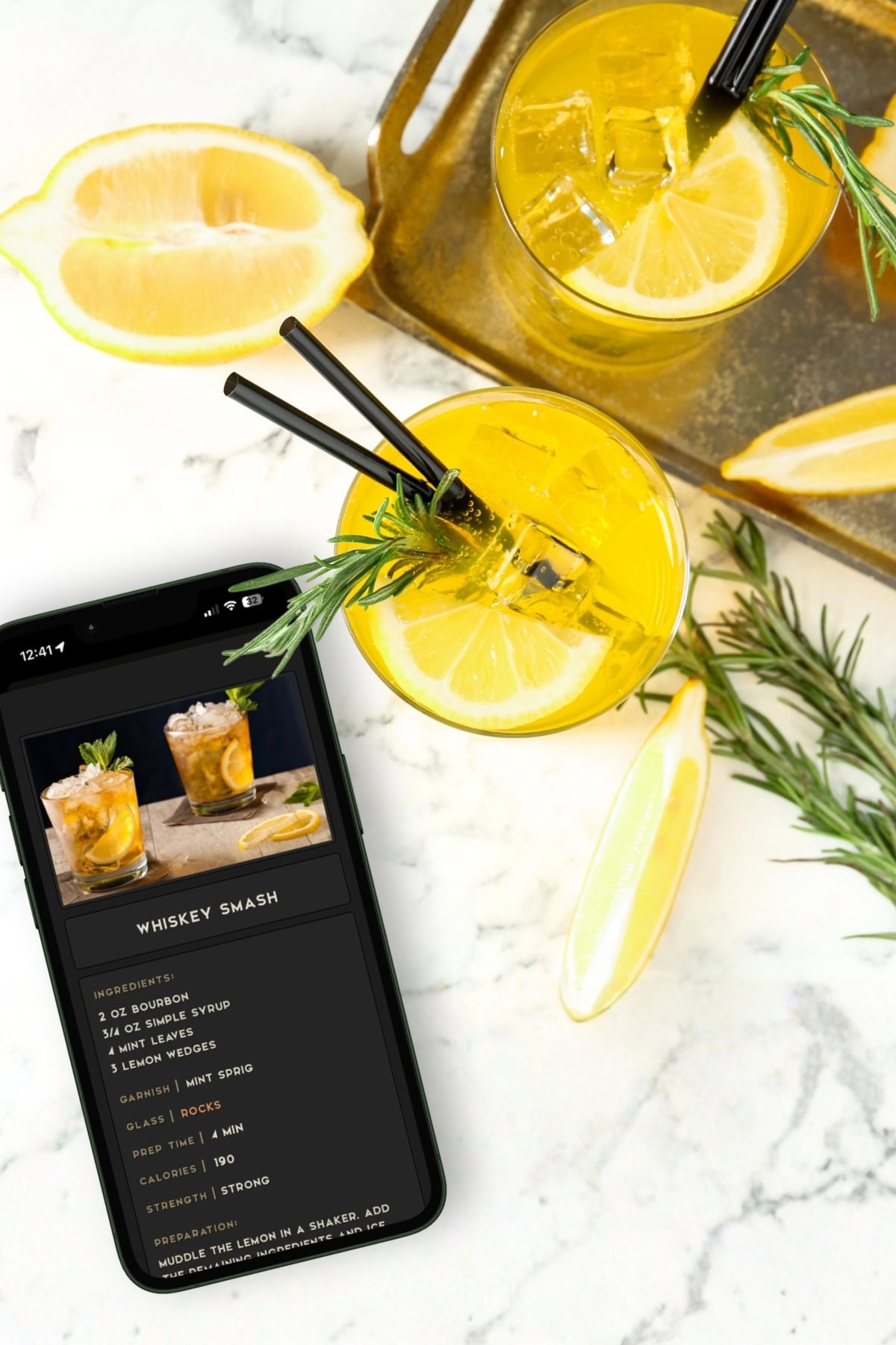 Overhead view of two whiskey smash cocktails with lemon slices and rosemary on a tray, next to a phone displaying the drink recipe, on a marble surface with lemon wedges.