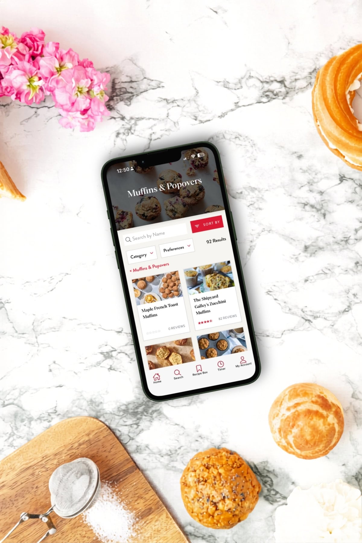 A smartphone displaying a baking recipe app featuring muffins and popovers rests on a marble surface surrounded by baked goods, a whisk, a sifter, powdered sugar, and pink flowers.