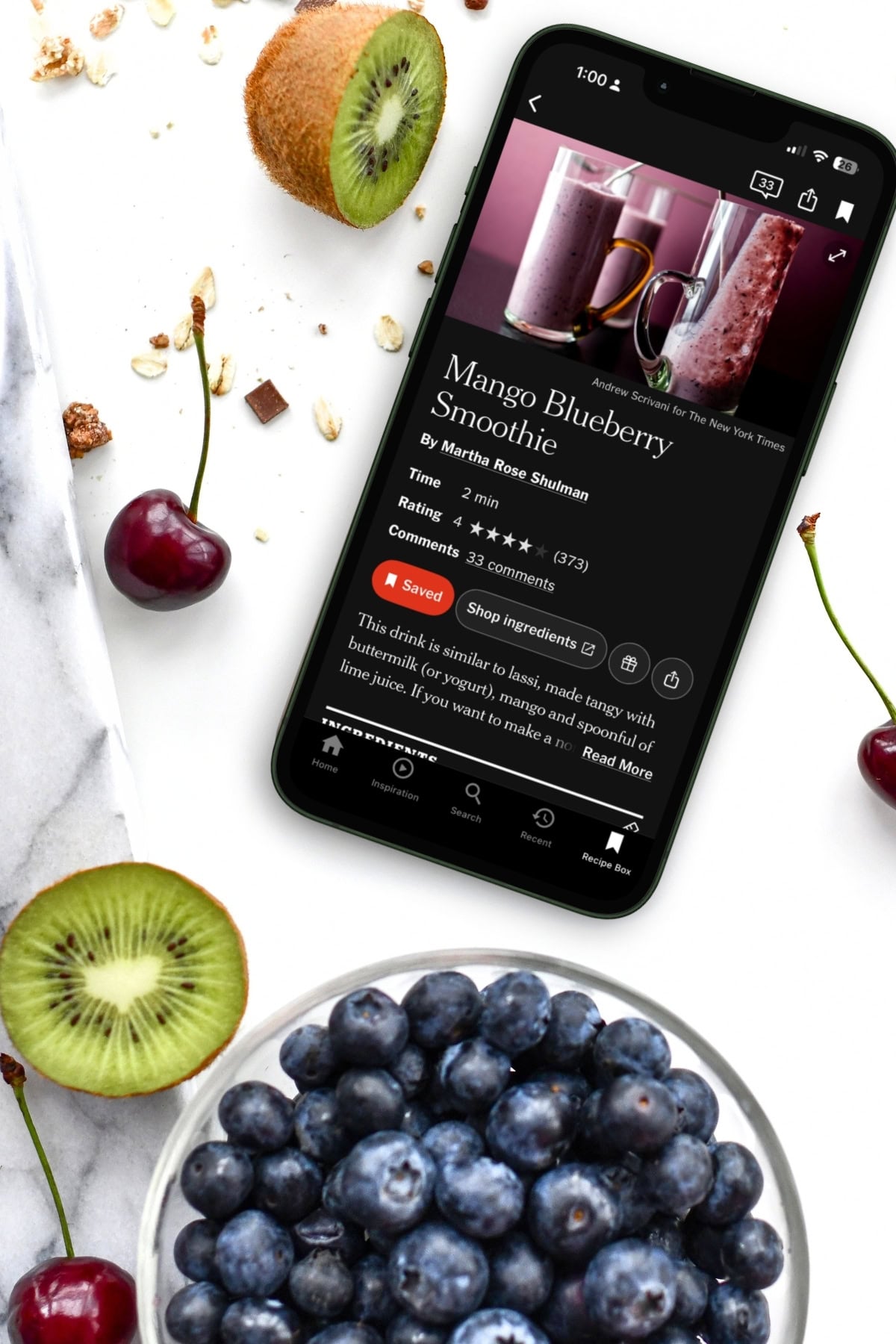A smartphone displays a Mango Blueberry Smoothie recipe on a marble surface, next to a bowl of blueberries, a halved kiwi, and scattered granola.