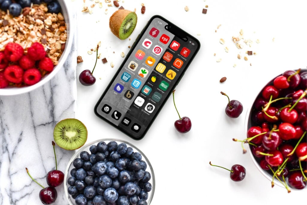A smartphone with colorful app icons lies on a white surface surrounded by bowls of raspberries, blueberries, cherries, granola, and kiwi slices.