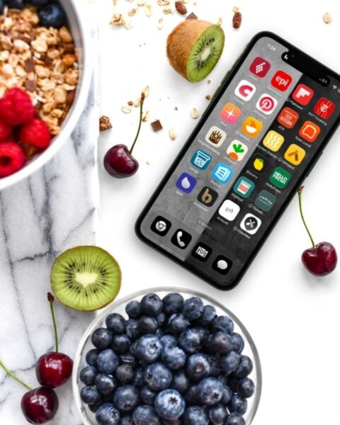 A smartphone with various app icons sits on a marble surface next to a bowl of blueberries, a bowl of granola with raspberries, cherries, and sliced kiwi.