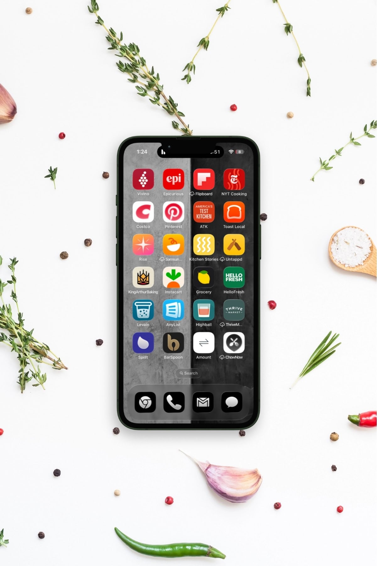 A smartphone displaying various cooking and food-related app icons on its screen, placed on a white surface with herbs, spices, and vegetables scattered around.