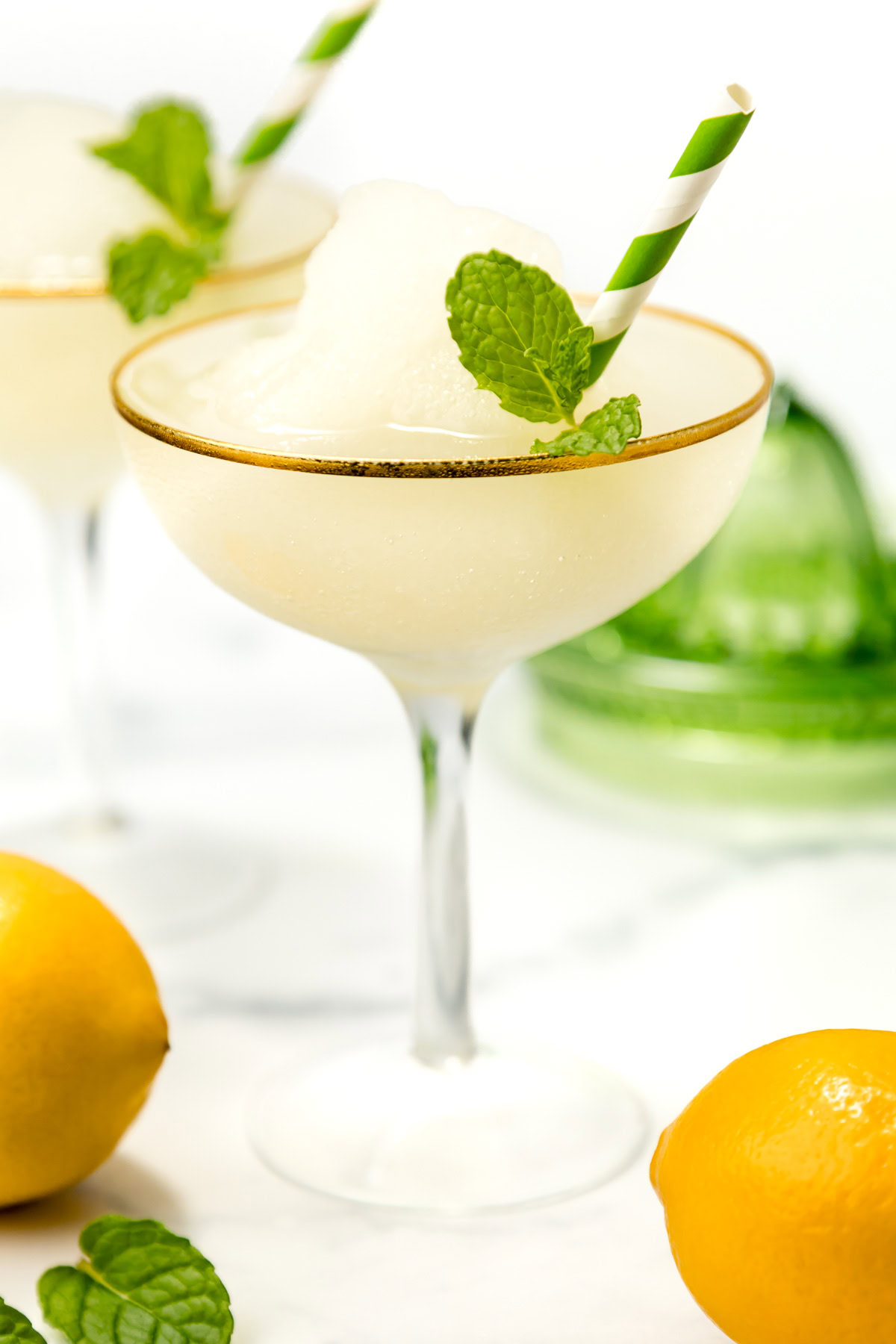 A frozen cocktail in a coupe glass with a green striped straw and mint garnish; lemons and mint leaves are nearby.