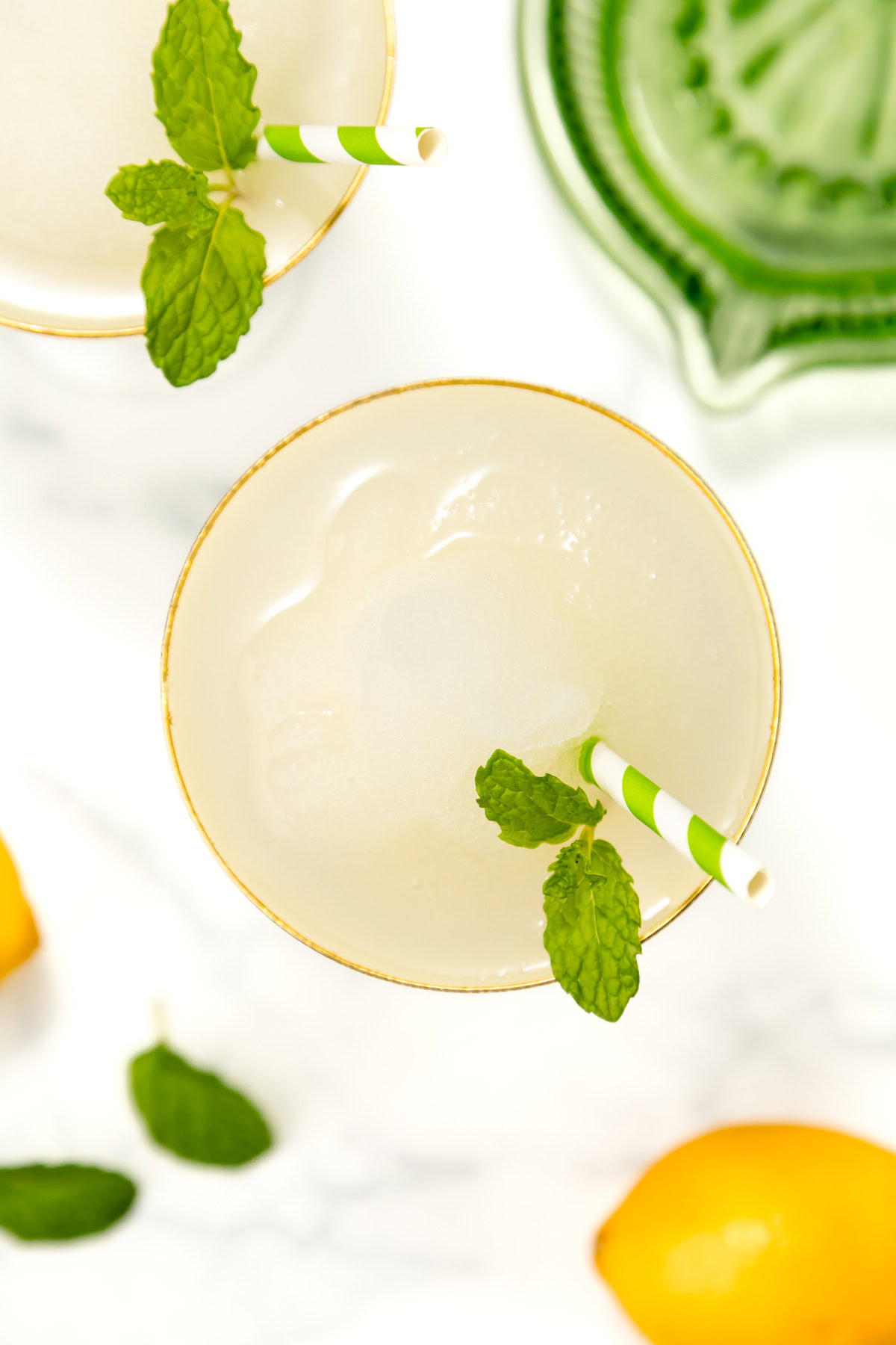 Overhead view of a glass of lemonade with a striped straw, ice, and mint garnish. Lemons, mint leaves, and a green citrus juicer are nearby on a white surface.