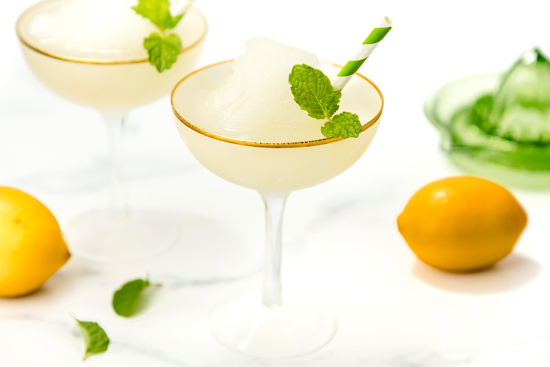 A frozen lemon cocktail in a coupe glass with a green striped straw and mint garnish, surrounded by lemons and mint leaves.