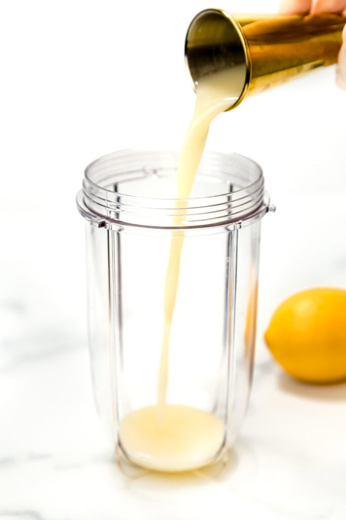 Lemon juice is being poured from a small metal cup into a clear blender cup, with a whole lemon on the side.