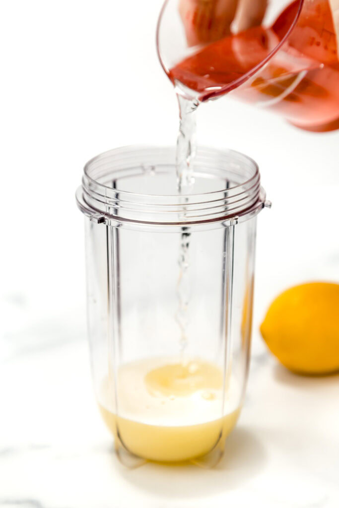 A clear blender cup with lemon juice inside is being filled with water from a glass measuring cup; a whole lemon sits nearby.