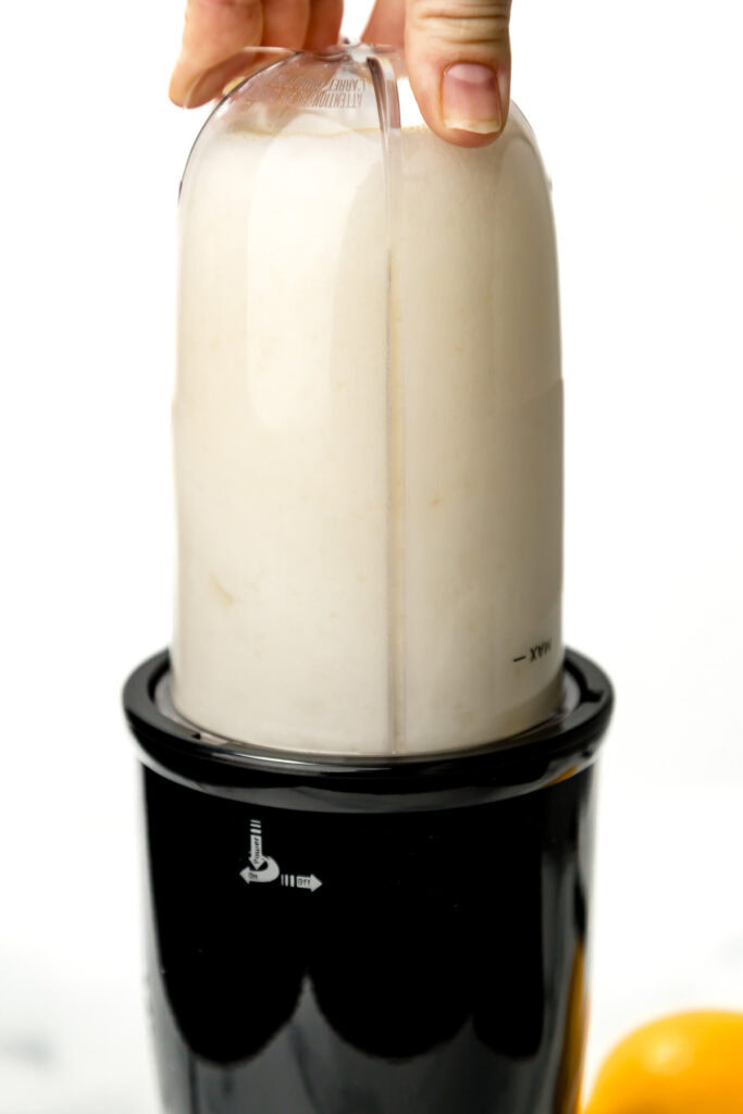 A hand holds the lid of a small countertop blender filled with a white liquid mixture, blending on a white background.