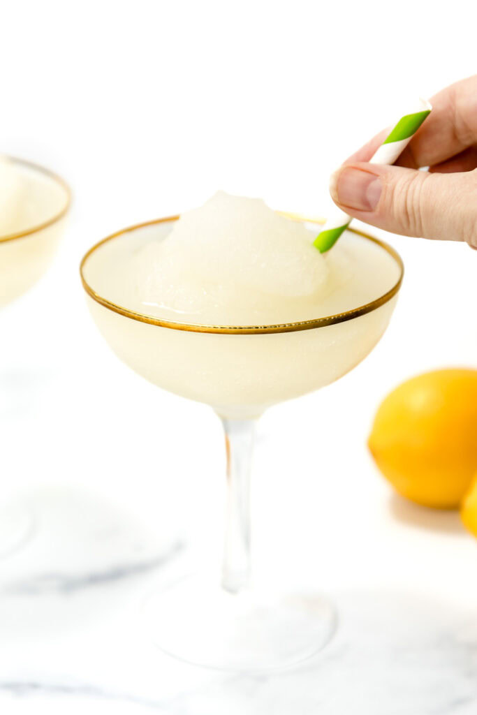 A hand holds a green and white striped straw, stirring a frozen lemon cocktail in a gold-rimmed glass with whole lemons in the background.