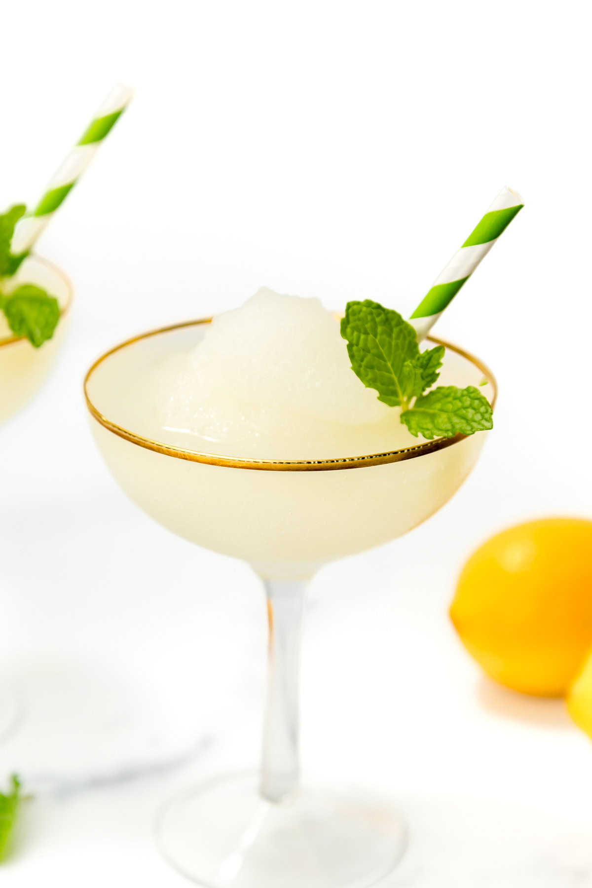 A coupe glass with a frozen white cocktail, garnished with a mint sprig and a green-striped straw, sits next to a lemon on a white background.