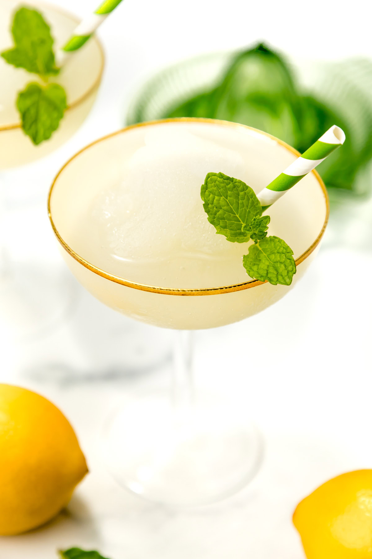 A cocktail glass with a frozen drink, garnished with a mint sprig and green-striped straw. Lemons and a green cloth are in the background.