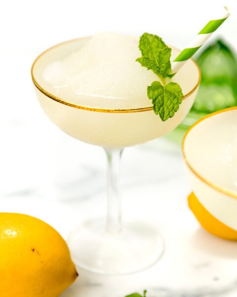 A frozen cocktail in a coupe glass garnished with a mint sprig and striped straw, next to a lemon and mint leaves on a white surface.