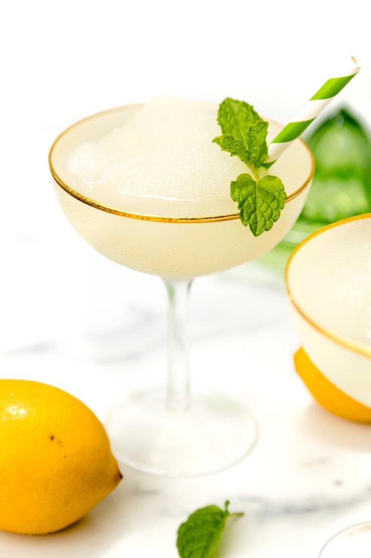 A frozen cocktail in a coupe glass garnished with a mint sprig and striped straw, next to a lemon and mint leaves on a white surface.
