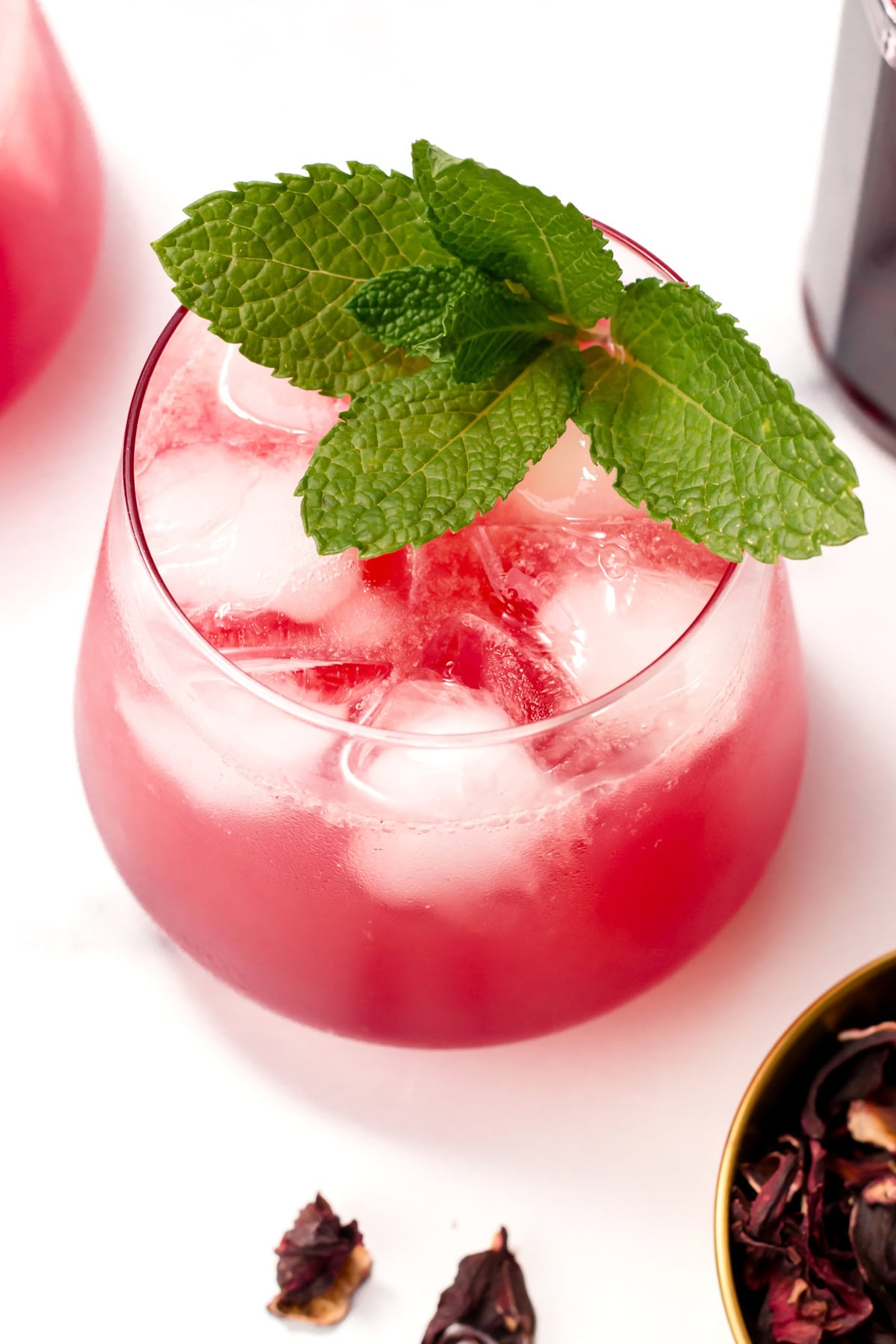 A glass of pink iced drink garnished with a sprig of fresh mint, with hibiscus petals and a brass bowl nearby on a white surface.