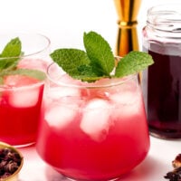 A glass of pink iced drink garnished with mint leaves sits on a white surface, with another glass, a gold jigger, a jar of dark liquid, and dried petals nearby.