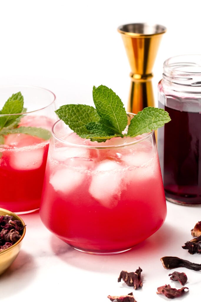 A glass of pink iced drink garnished with mint leaves sits on a white surface, with another glass, a gold jigger, a jar of dark liquid, and dried petals nearby.