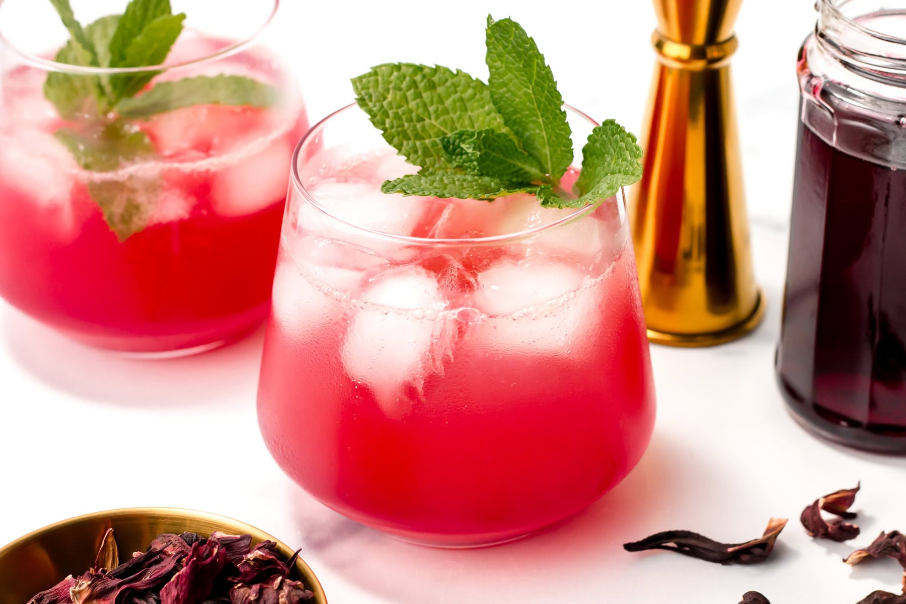Two glasses of iced red hibiscus drink garnished with mint leaves, with a jar of dark liquid, a brass jigger, and dried hibiscus petals nearby on a white surface.