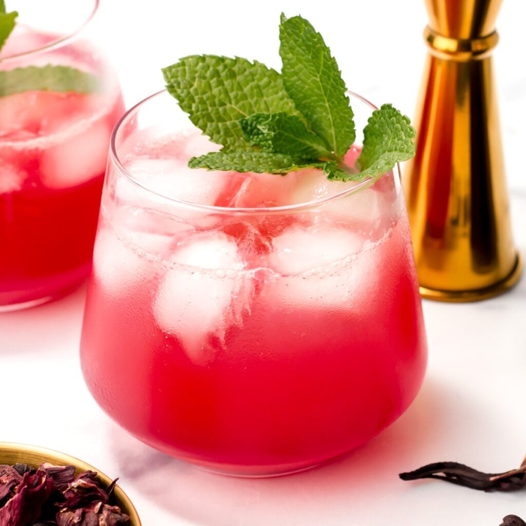 A glass of pink iced drink garnished with fresh mint leaves, with another glass, a brass jigger, and dried hibiscus petals nearby on a white surface.