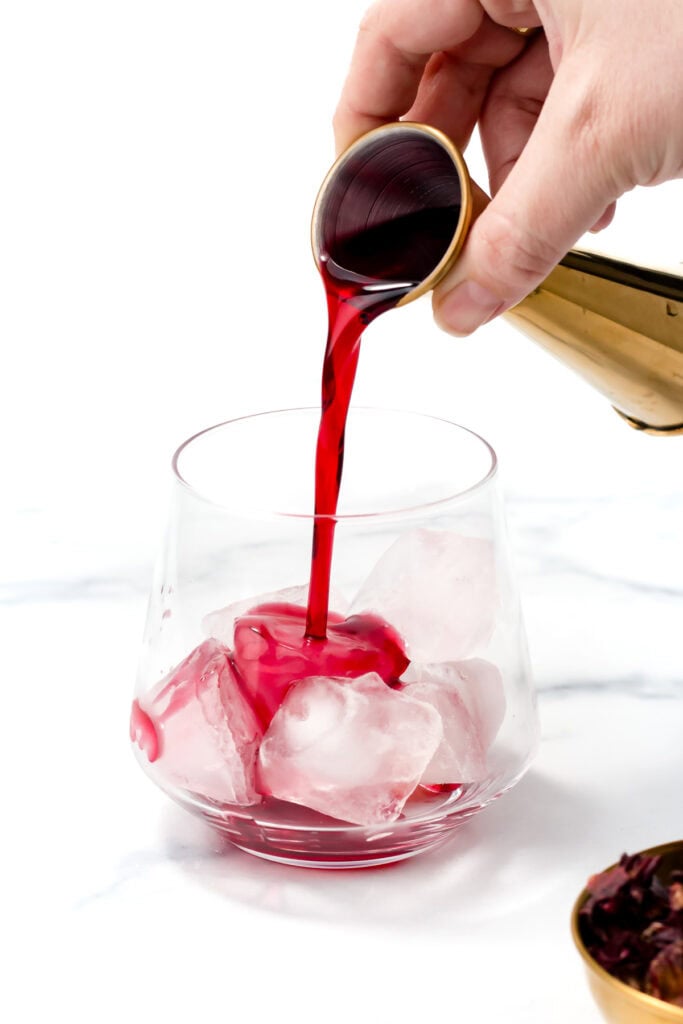 A hand pours a dark red liquid from a jigger into a glass filled with ice cubes on a white surface.